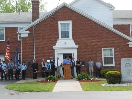 People in formal attire stand in front of a brick building; one person speaks at a podium on the steps.