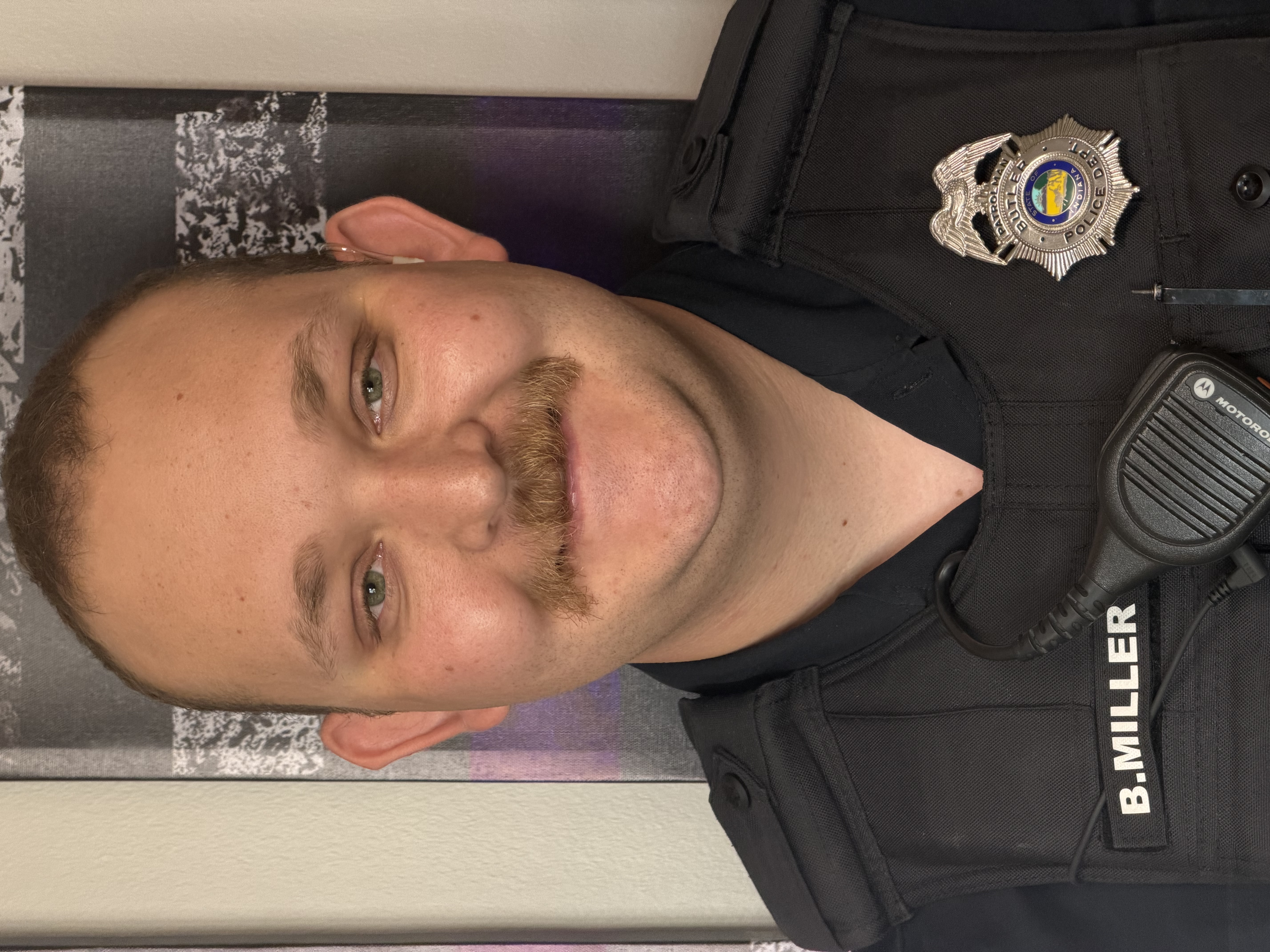 Close-up head-and-shoulders photo of a police officer in a dark uniform; badge and name tag read “B. MILLER.”