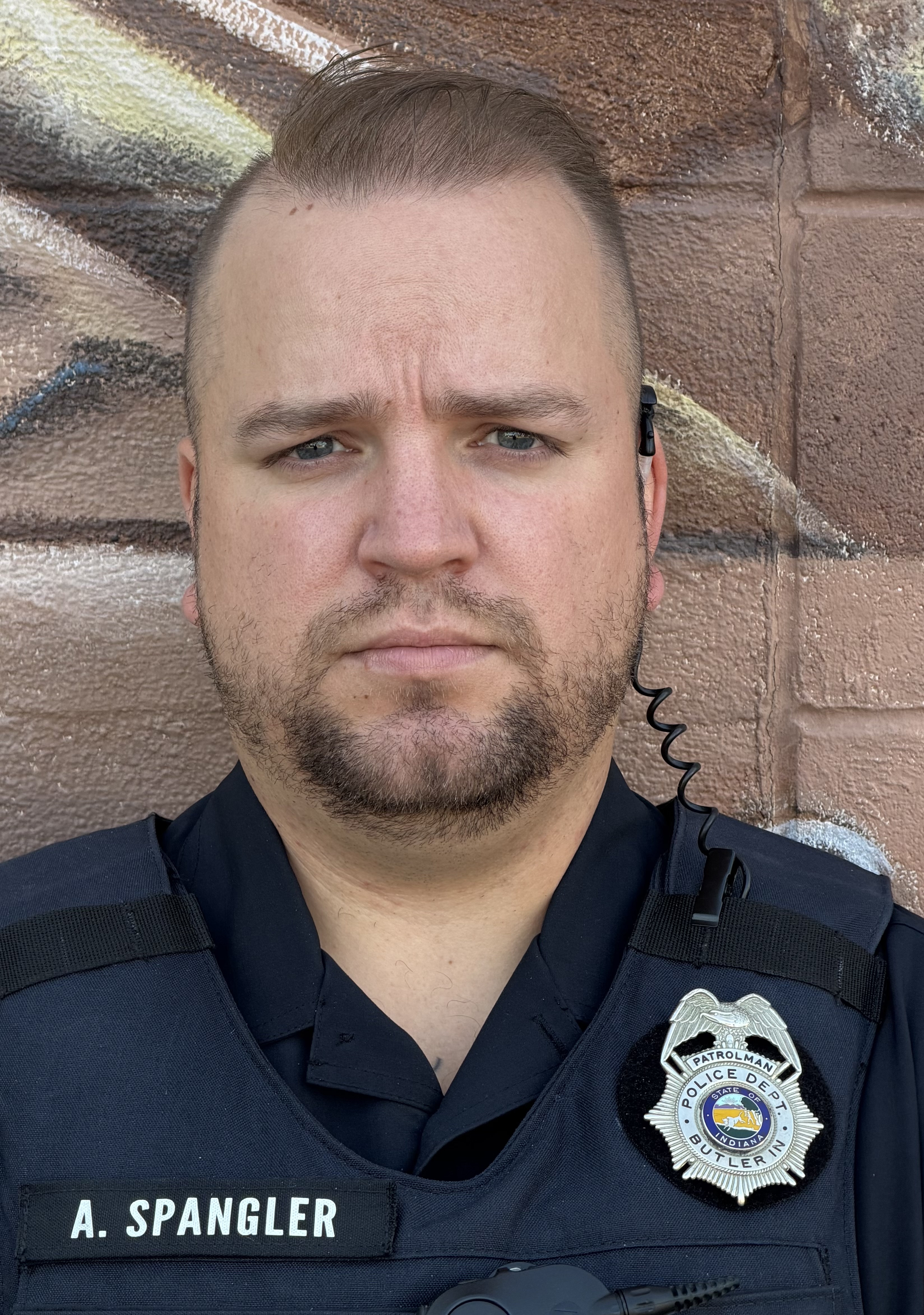 Uniformed officer in black with shoulder badge and name tag “A. SPANGLER,” photographed outdoors against a brick wall.