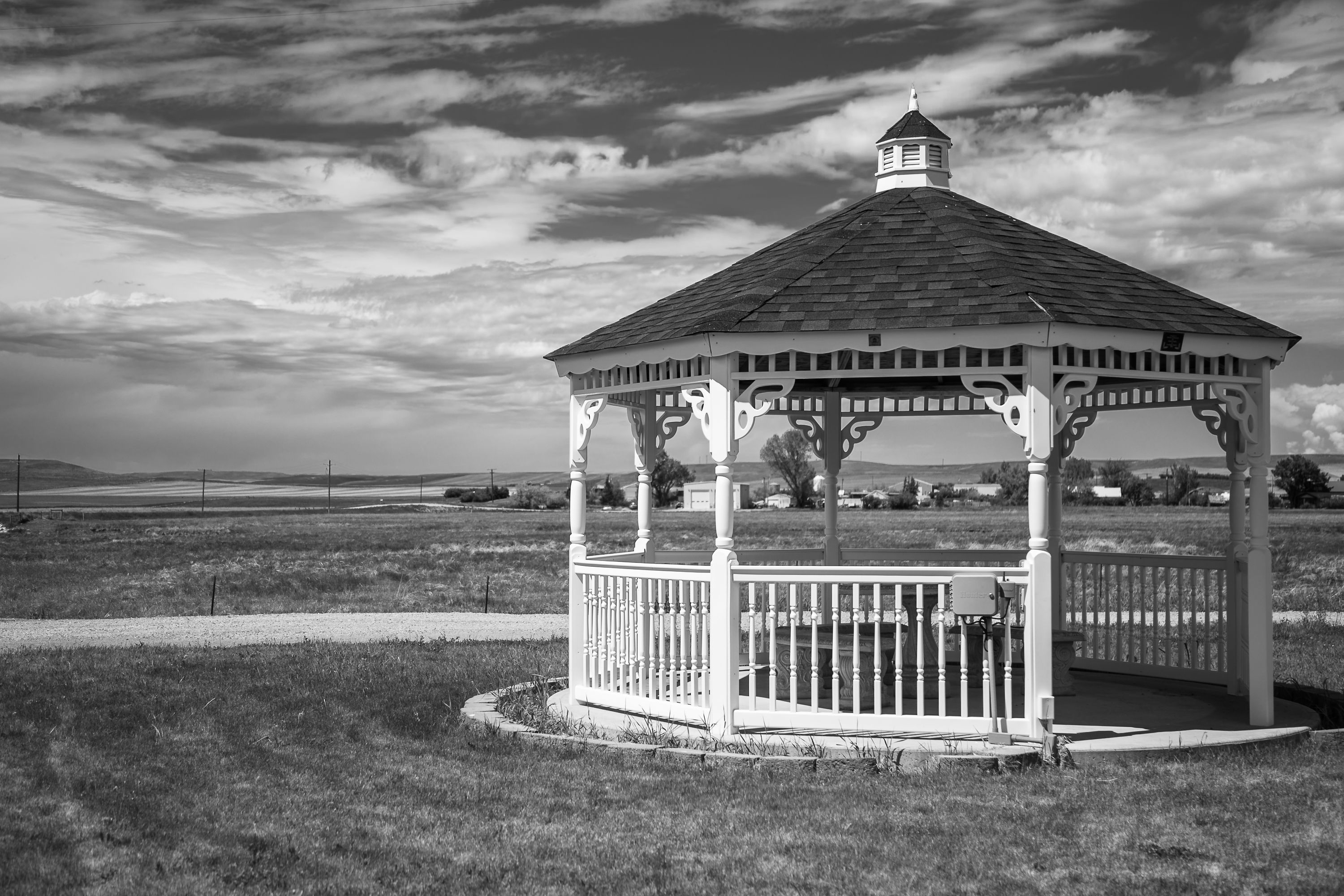 Black-and-white image of a white gazebo with a dark shingled roof in a grassy field under stormy clouds.