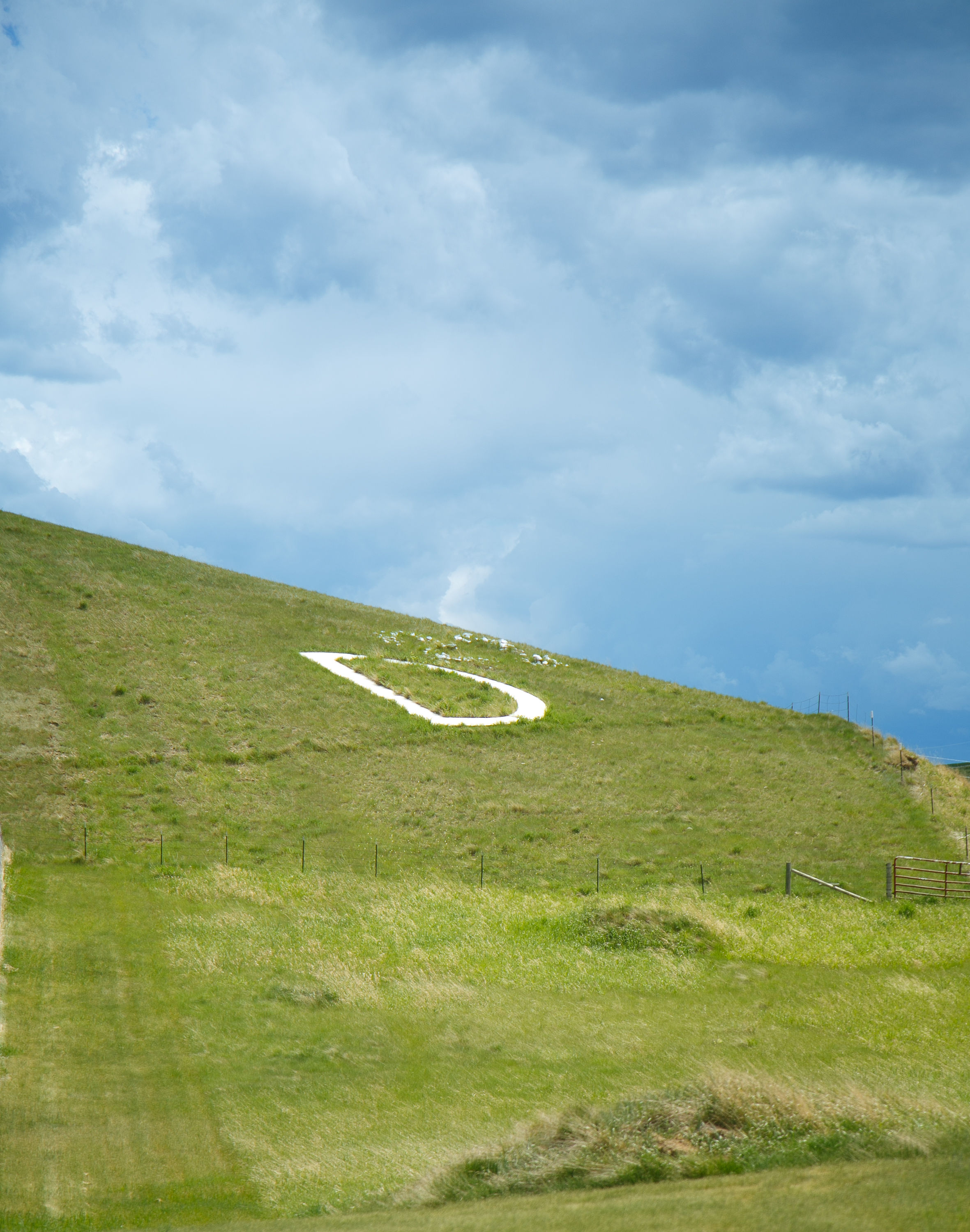Green grassy hillside with a narrow white curving path and low fencing under cloudy blue sky.