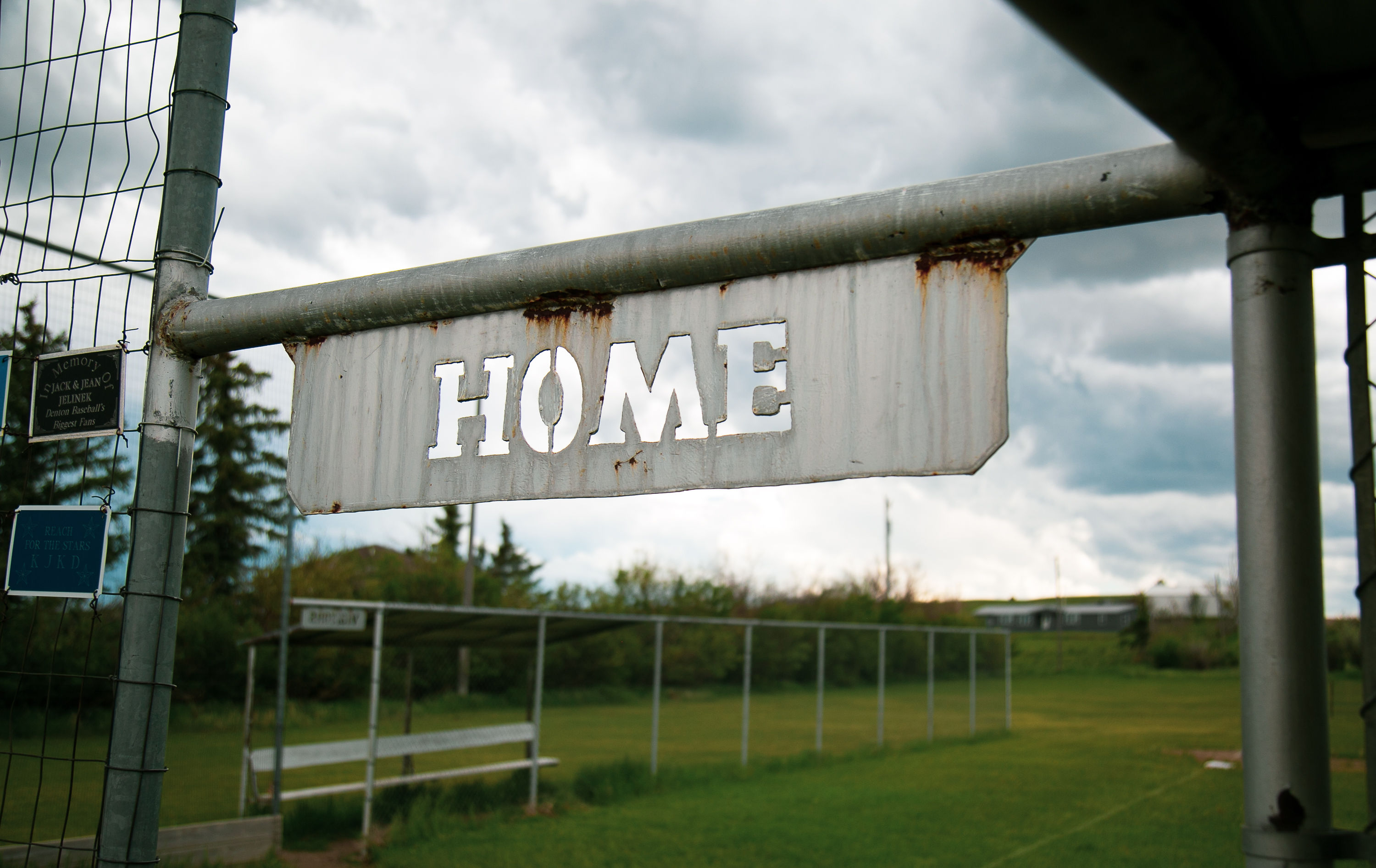 Fence sign on a metal beam reads “HOME,” with rust stains and weathering visible under gray sky.