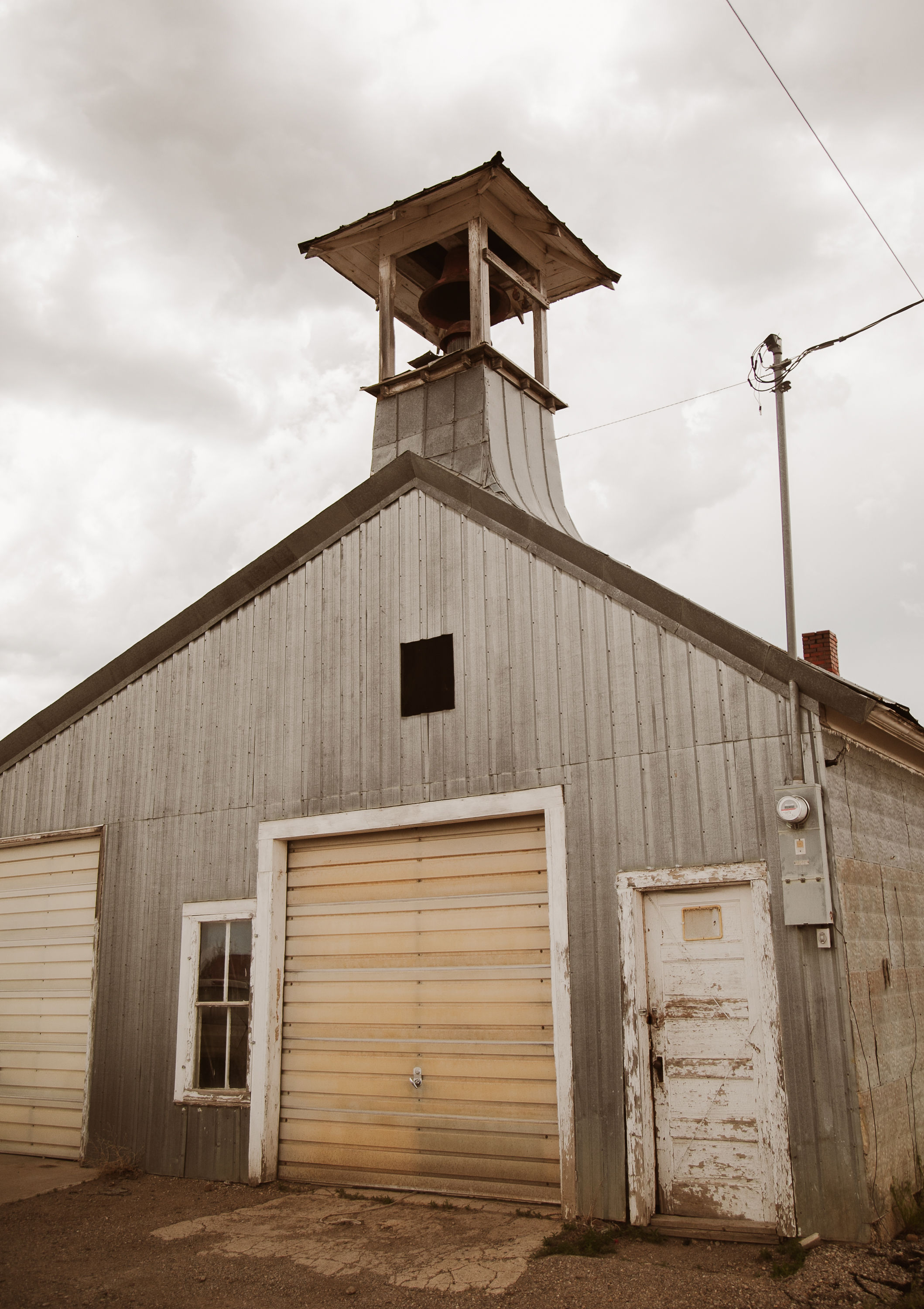Old wooden bell tower on a metal-sided building with damaged doors and overhead wiring in overcast sky.