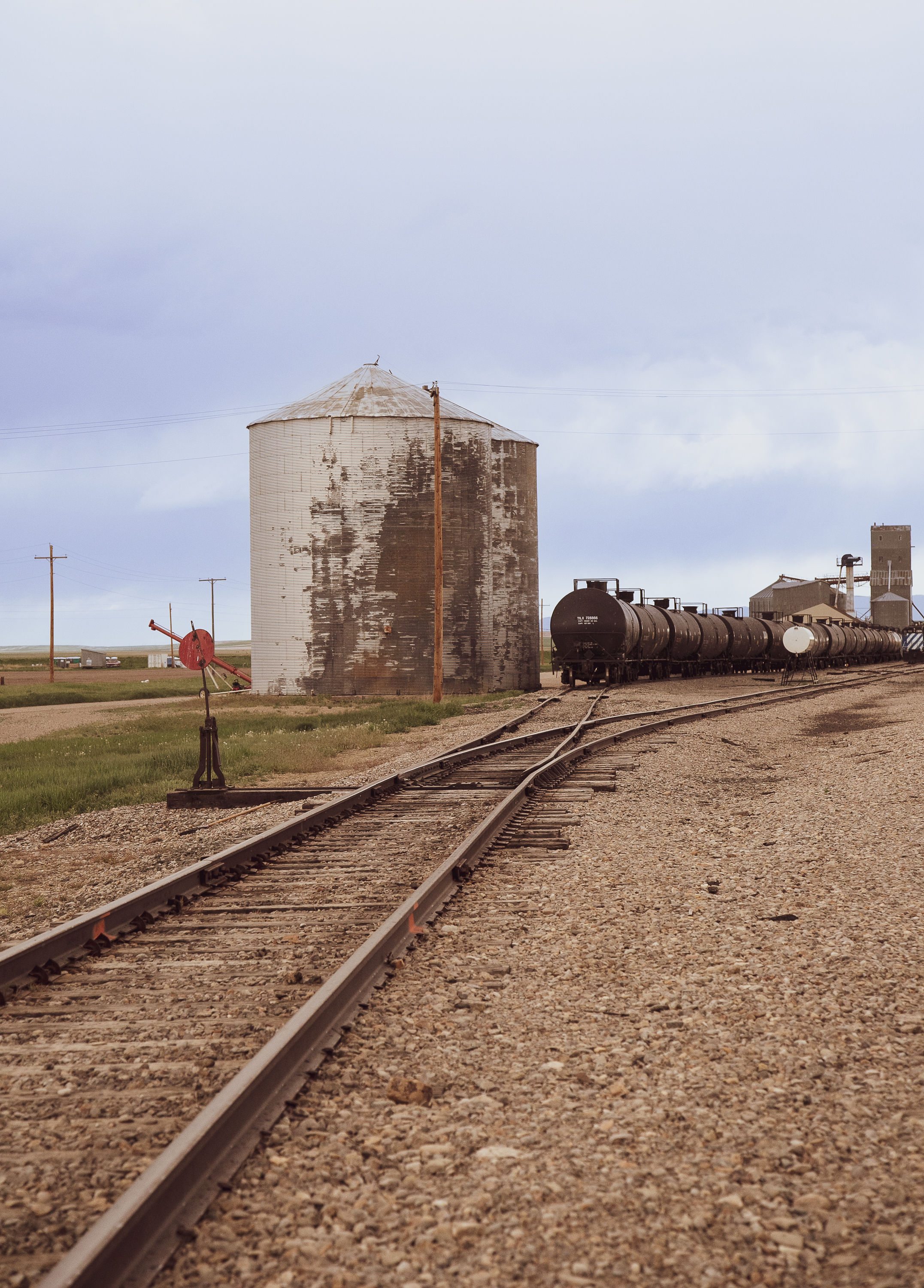 Weathered grain silo beside railroad tracks; freight tank cars and utility buildings in a rural landscape under cloudy sky.