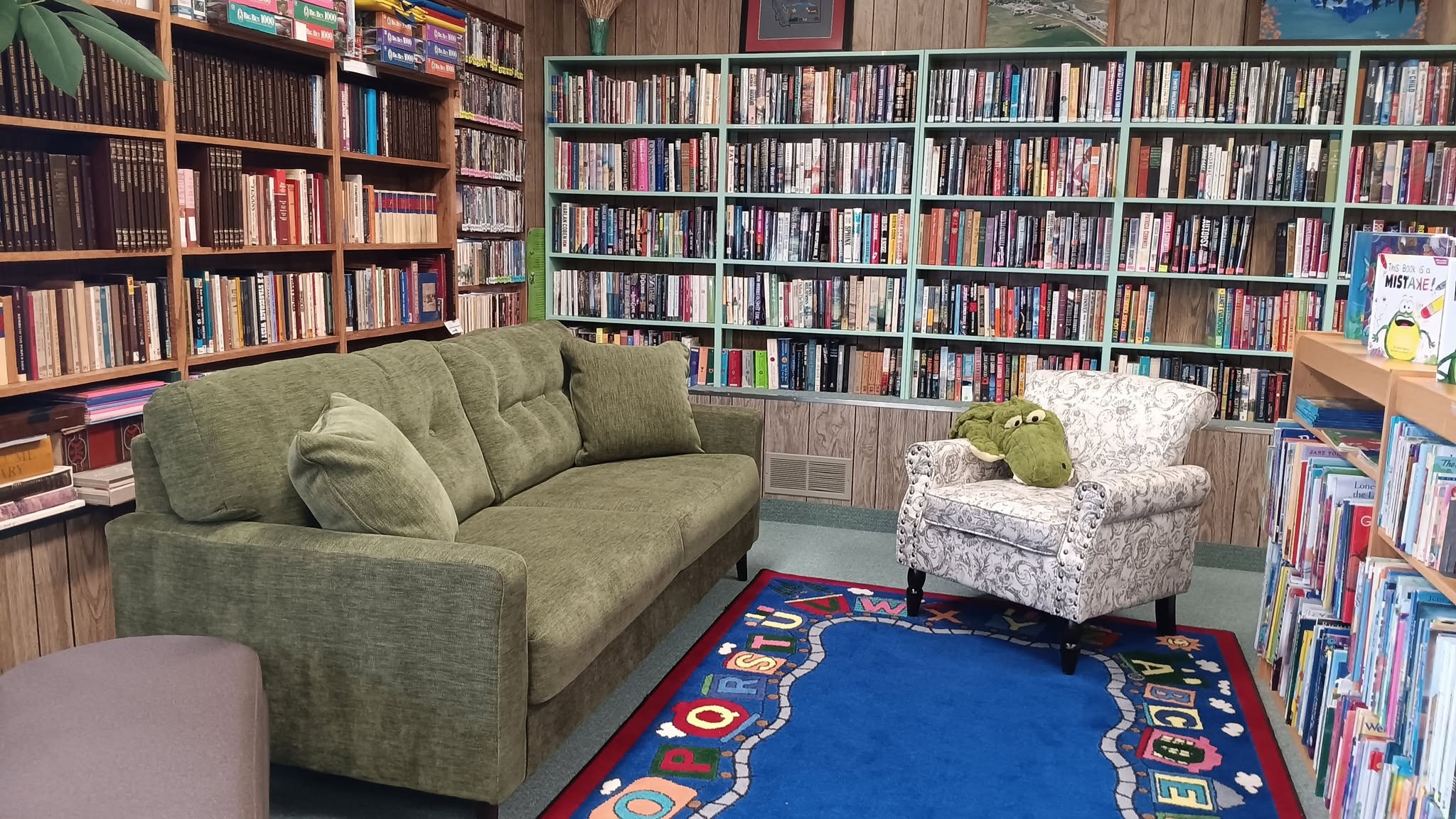 Library reading nook with bookshelves, green sofa, patterned armchair, and blue alphabet play mat.
