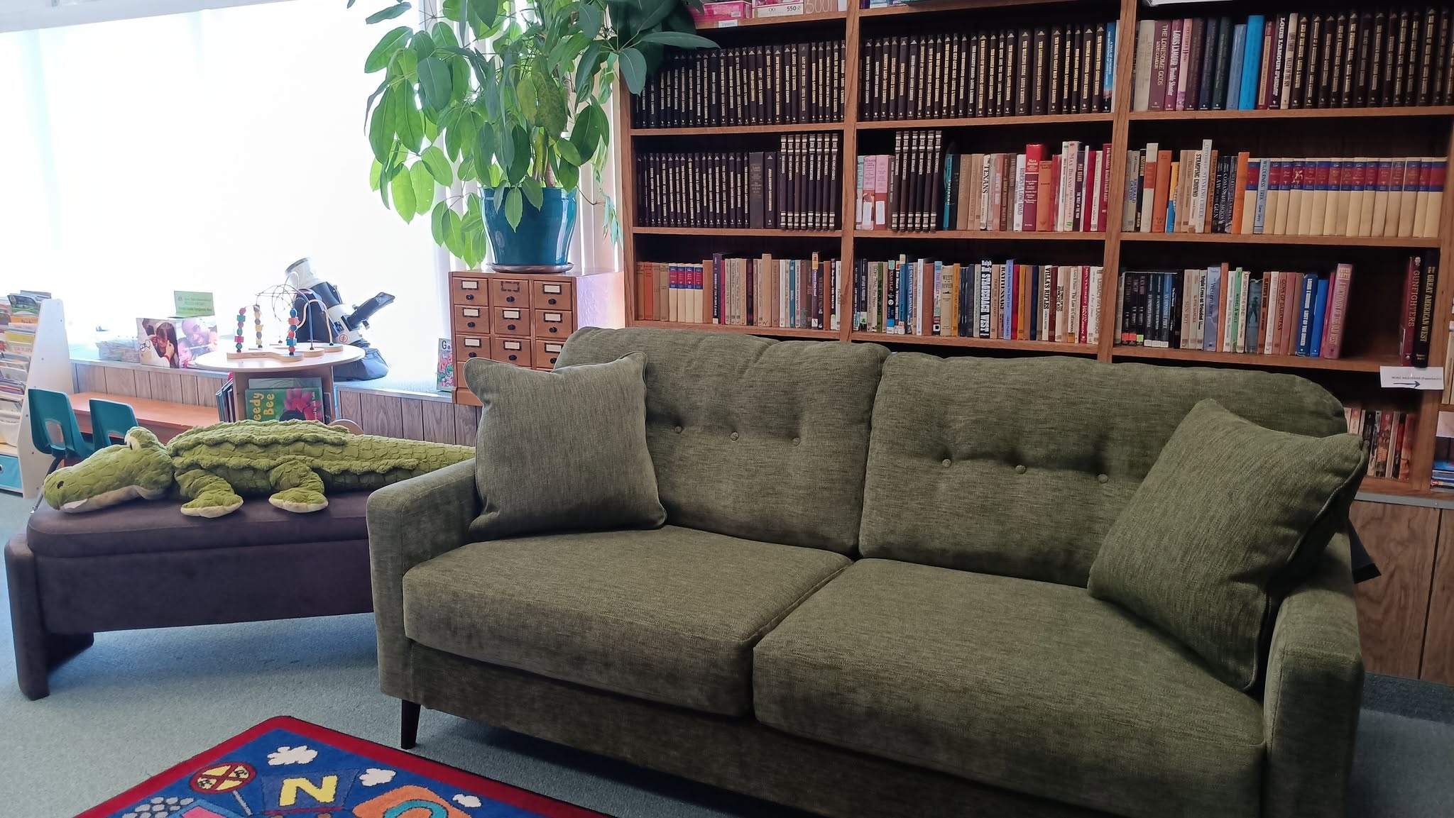 Green sofa and pillows in a children’s reading room, with tall wooden bookshelves and toy items nearby.