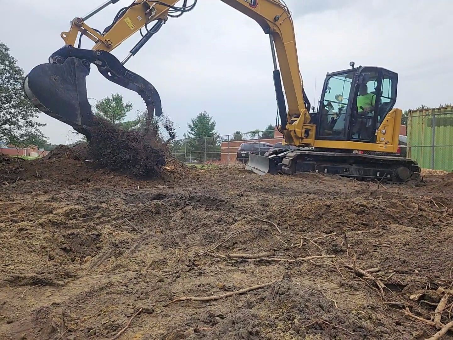 Yellow tracked excavator bucket digging into uneven soil; soil/roots scattered around work area.