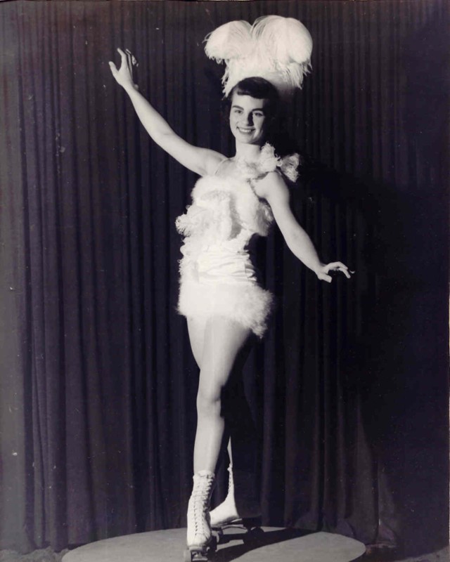Black-and-white photo of a showgirl in a feathered costume posing with one arm raised before a dark curtain.