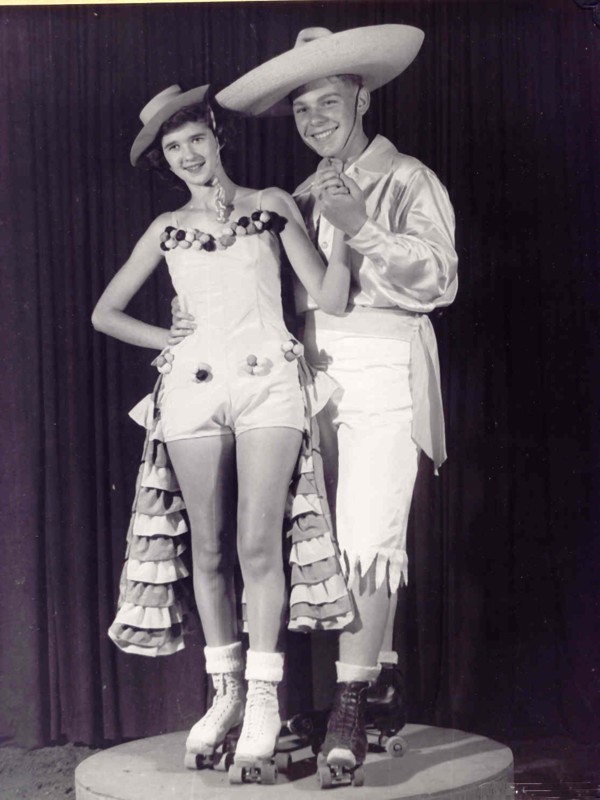 Two people in white cowboy-style outfits pose on roller skates in front of a dark curtain.