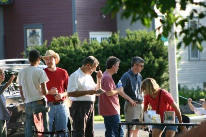 People chatting and browsing items at an outdoor yard sale in front of houses.