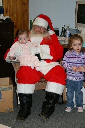 Santa in red suit and hat sits indoors holding a baby; two children stand nearby.