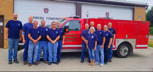 Group of firefighters in blue shirts standing in front of a red fire truck outside a fire station.