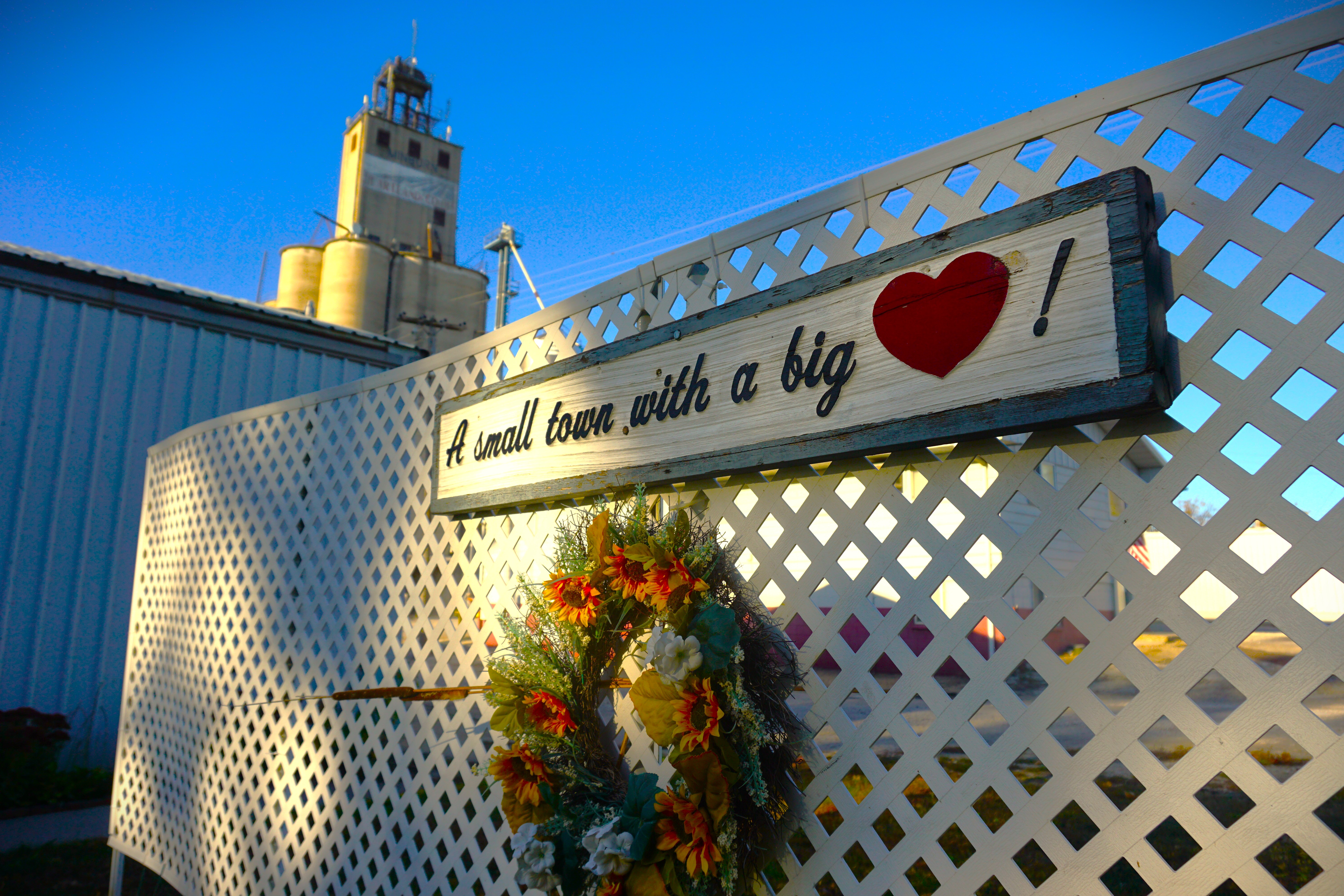 Wooden sign on a lattice fence reading “A small town with a big heart,” with a red heart and flowers below.