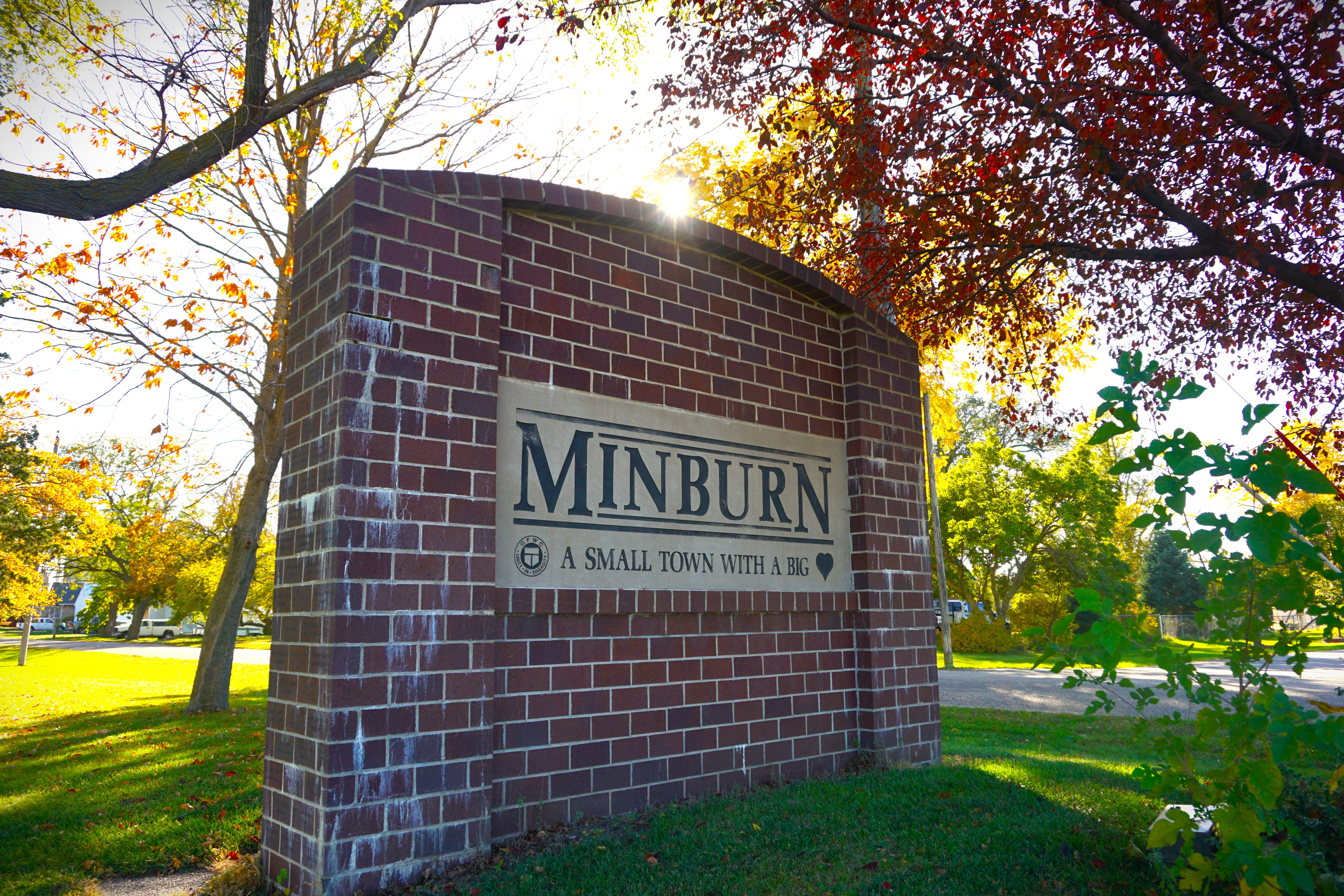 Brick entrance monument sign reading “MINBURN” and “A SMALL TOWN WITH A BIG” with autumn trees and sunlight behind it.