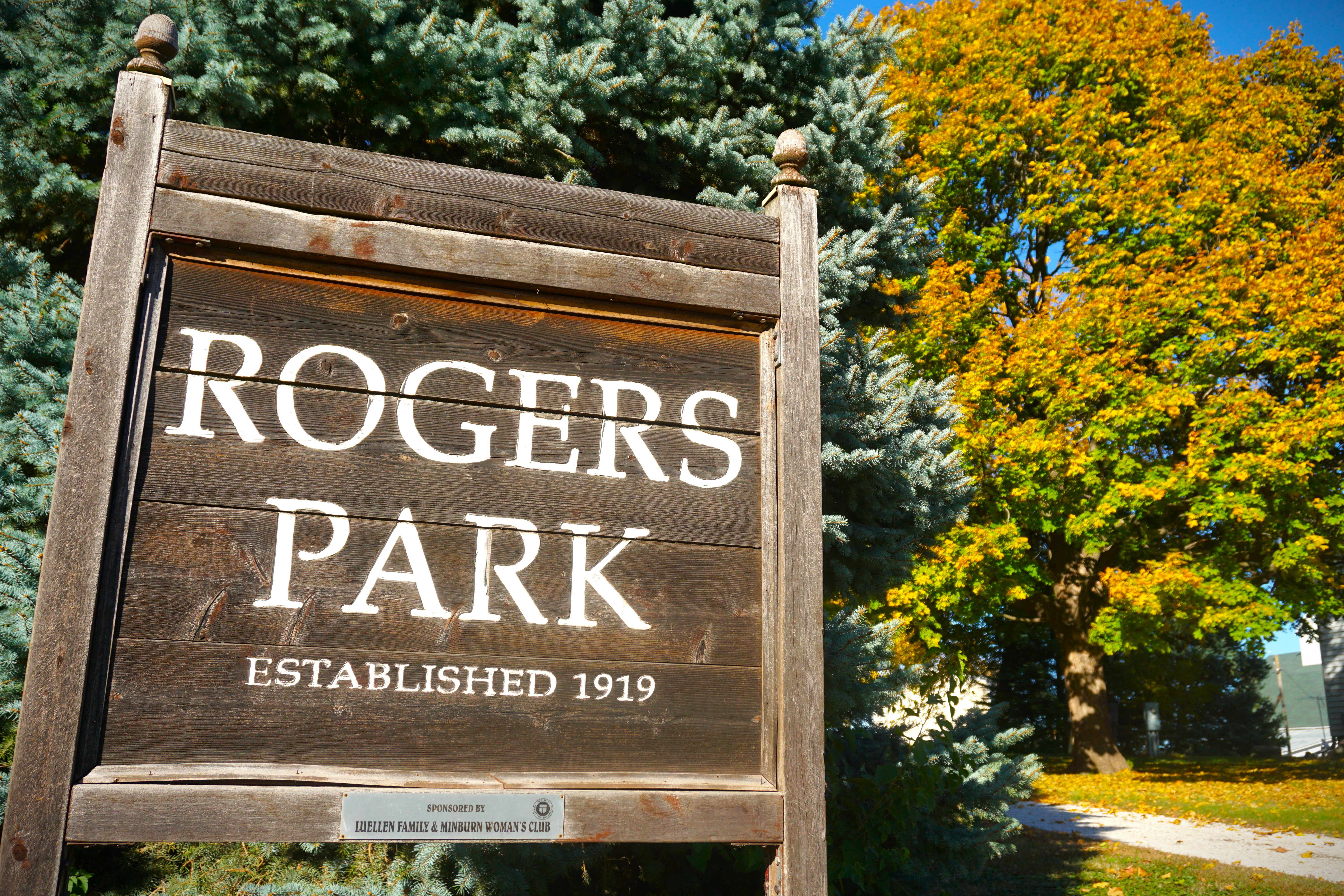 Wooden sign with white text: “Rogers Park,” “Established 1919,” in front of evergreen and yellow autumn trees.