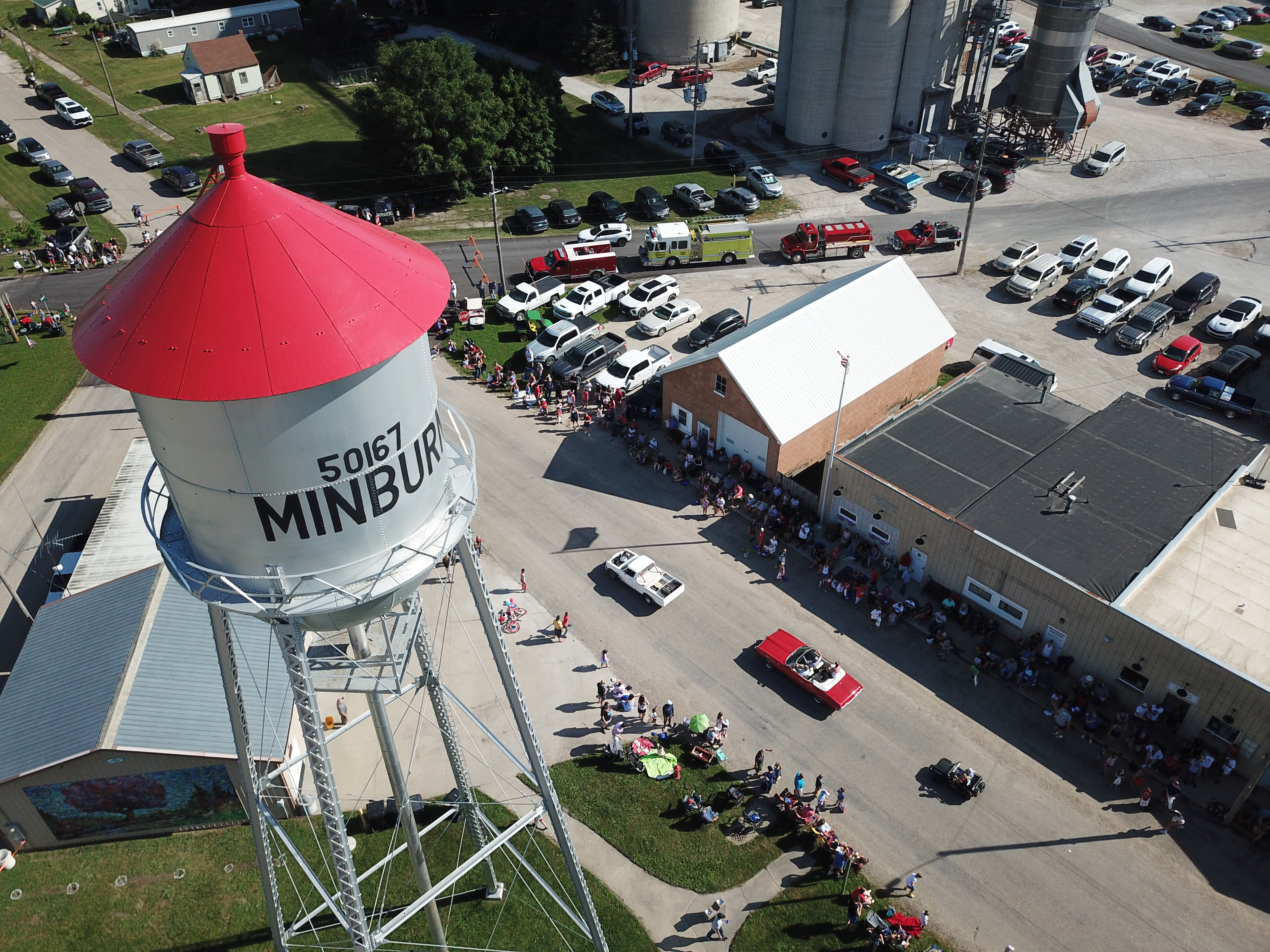 Aerial view of a crowd near a building and parking lot with fire and EMS vehicles and trucks parked, including a large water-tower structure.