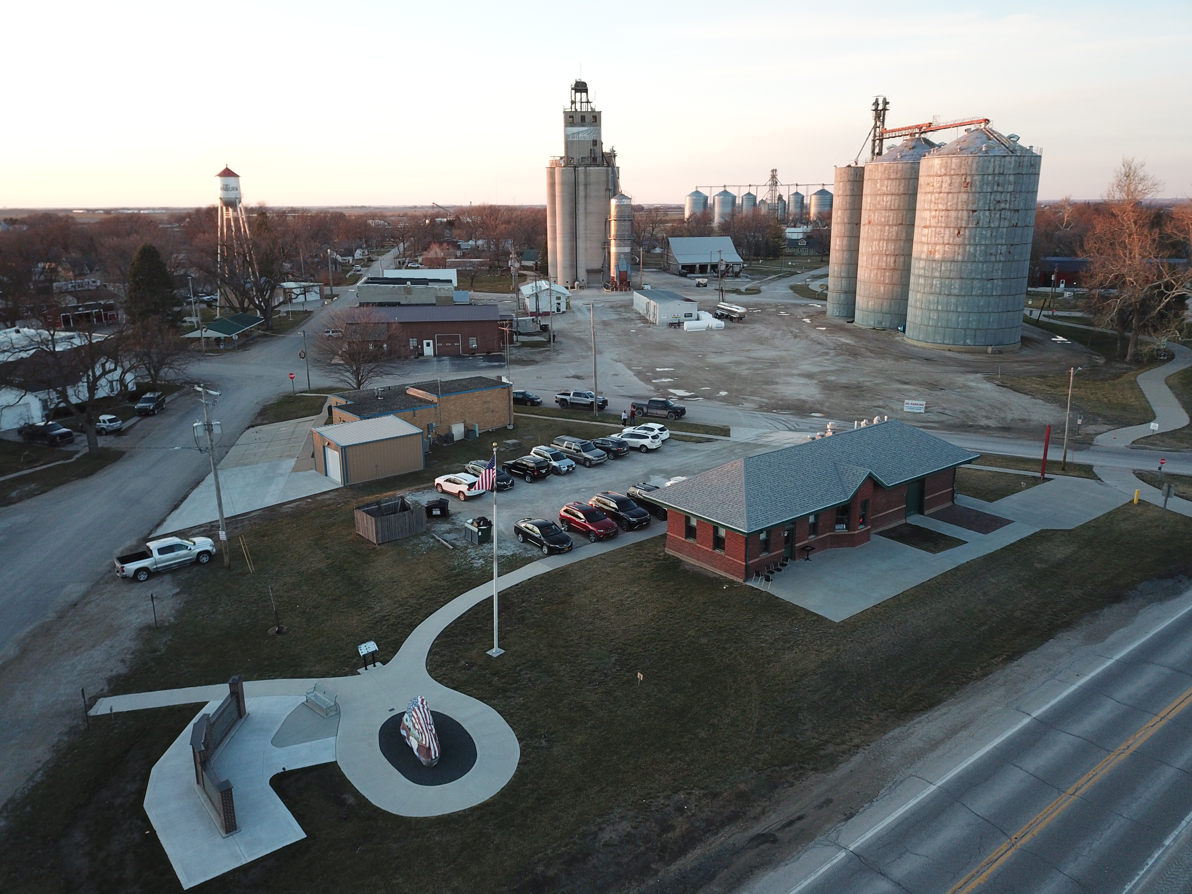 Aerial view of an industrial facility with tall silos and storage tanks, service roads, parking area, and nearby buildings.