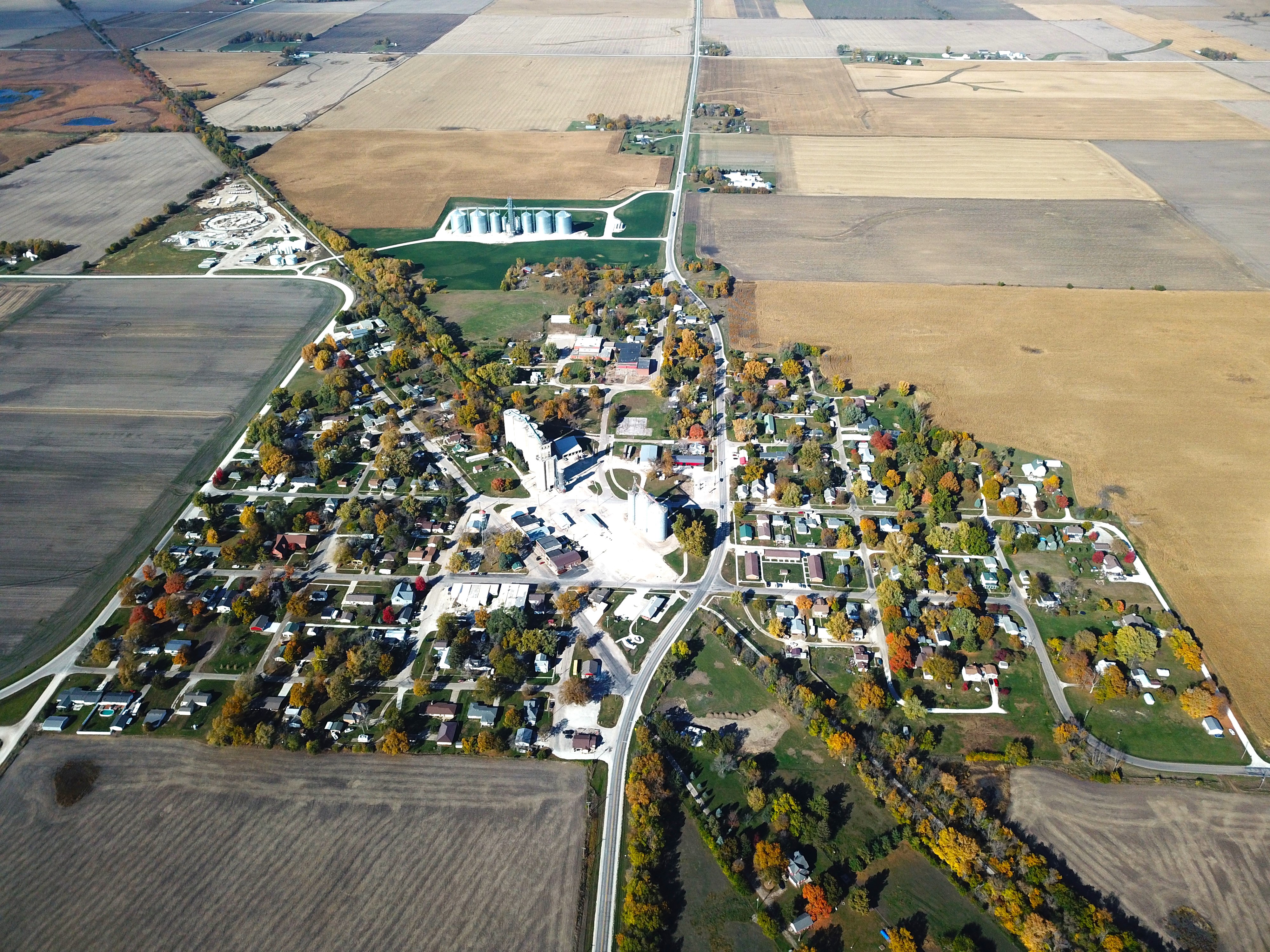 Aerial view of a rural town with many houses and roads, surrounded by farmland; a small industrial/agricultural site is visible near the center-left.