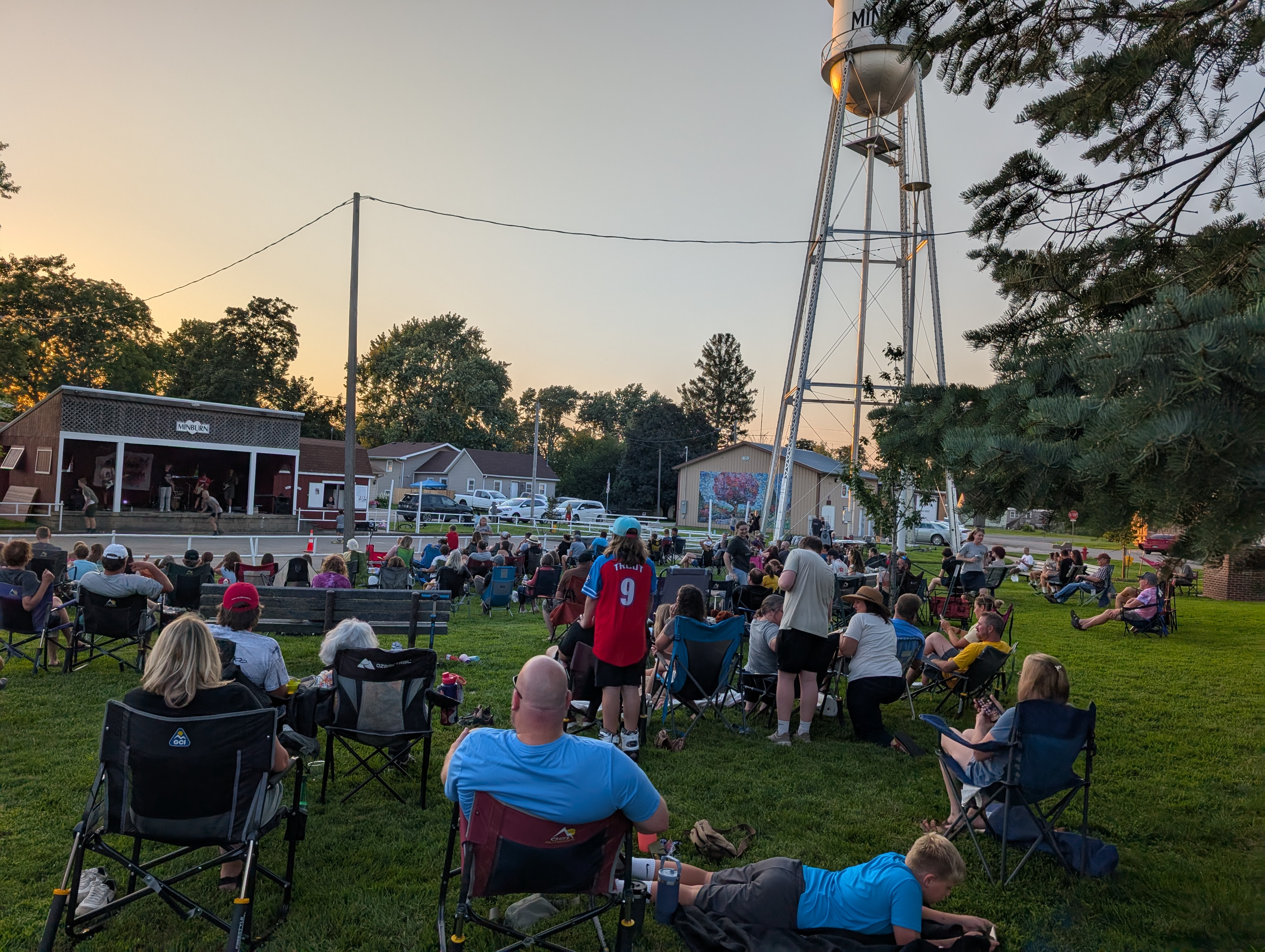 People seated on lawn chairs at an outdoor event near a small stage building; tall light tower overhead.