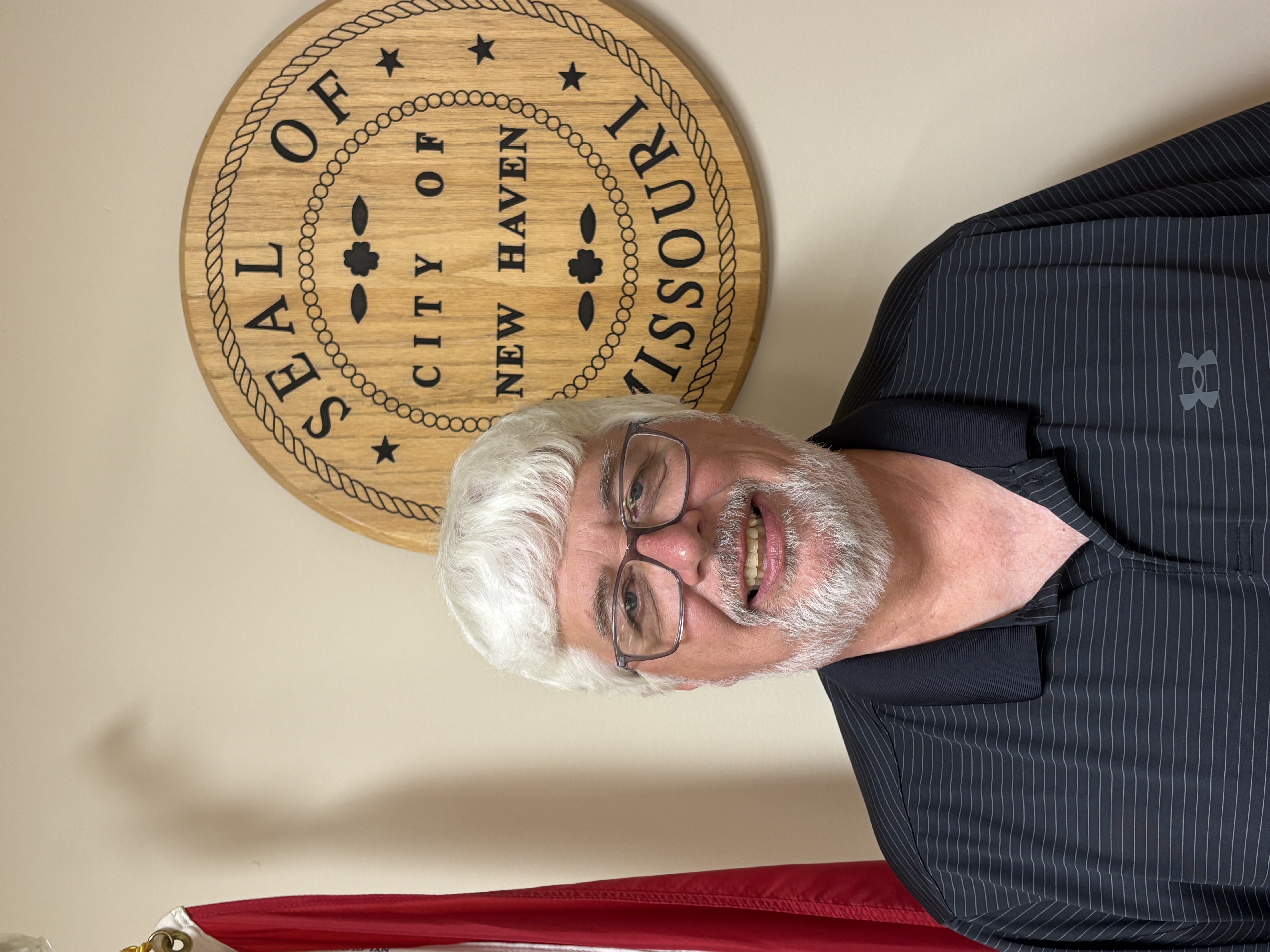 Man in striped polo shirt smiling in front of a wooden seal reading “Seal City of New Haven” with small stars and rope border.