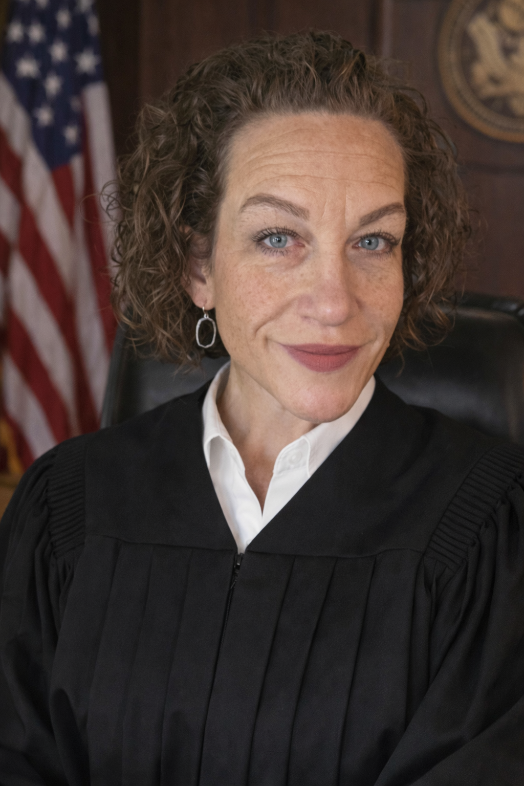 Woman in black judicial robe with white collar, wearing drop earrings, seated in front of U.S. flag.