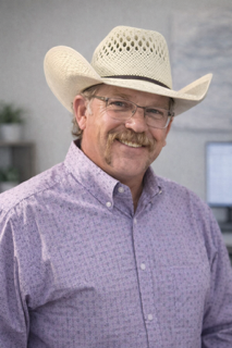 Smiling older man wearing a white cowboy hat and purple button-up shirt.