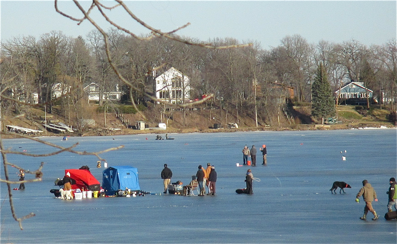 People ice fishing on a frozen Eagle Lake with portable shelters, a dog, and houses along the snowy shoreline