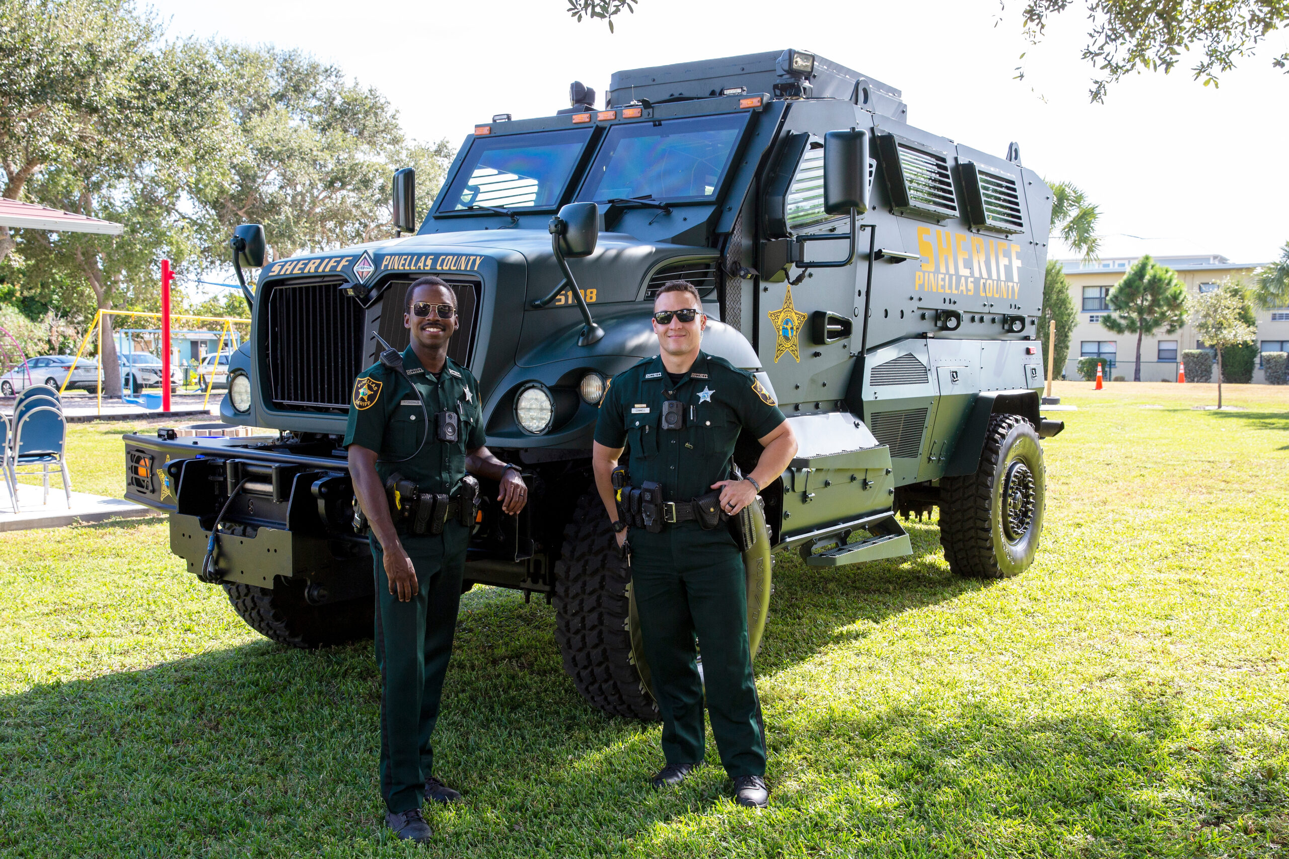 Two sheriff’s officers in uniform pose in front of a Pinellas County armored vehicle on grass.