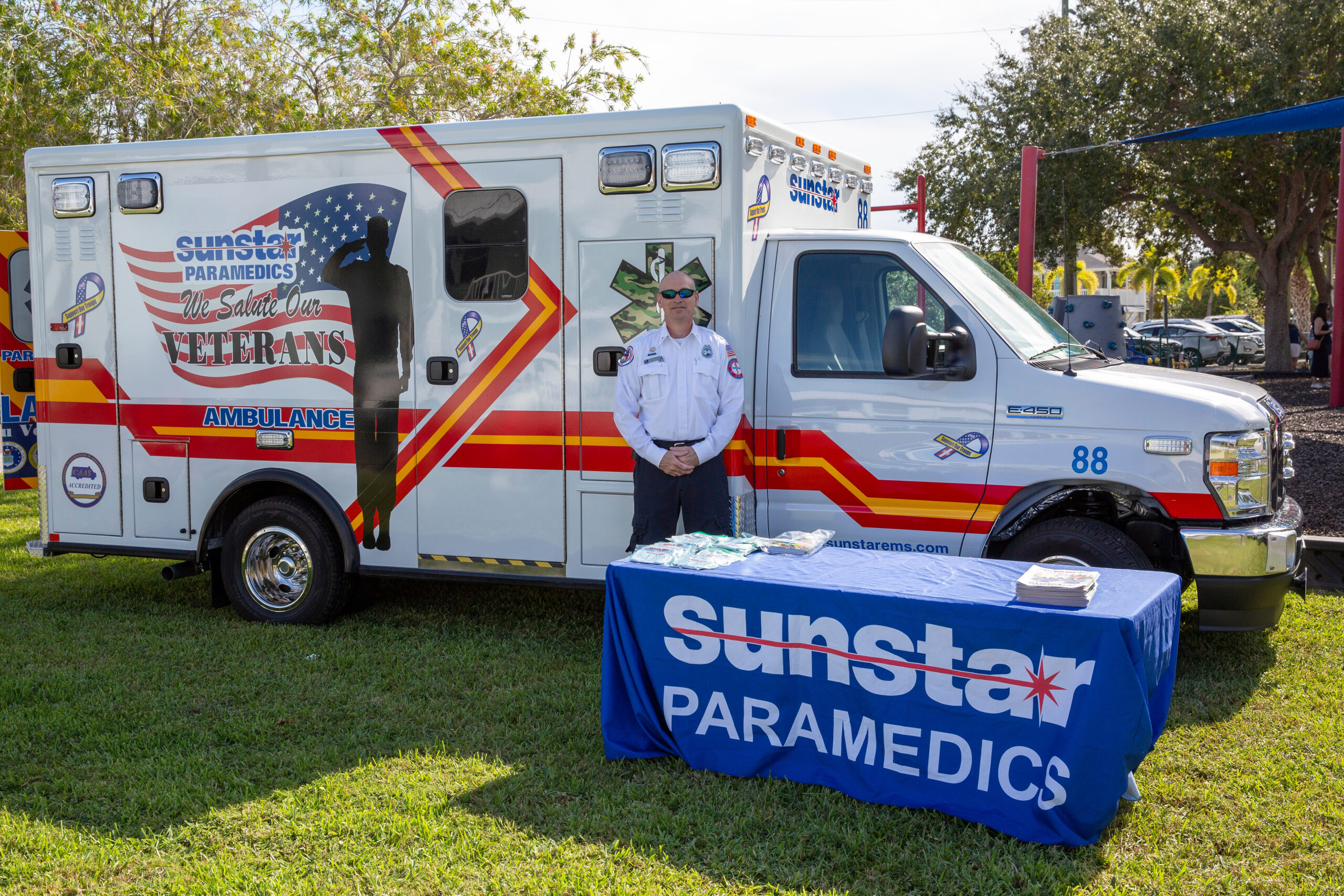 Paramedic stands beside white ambulance with “Sunst ar Paramedics” and “We Salute Our Veterans” graphics; table sign reads “Sunstar Paramedics.”