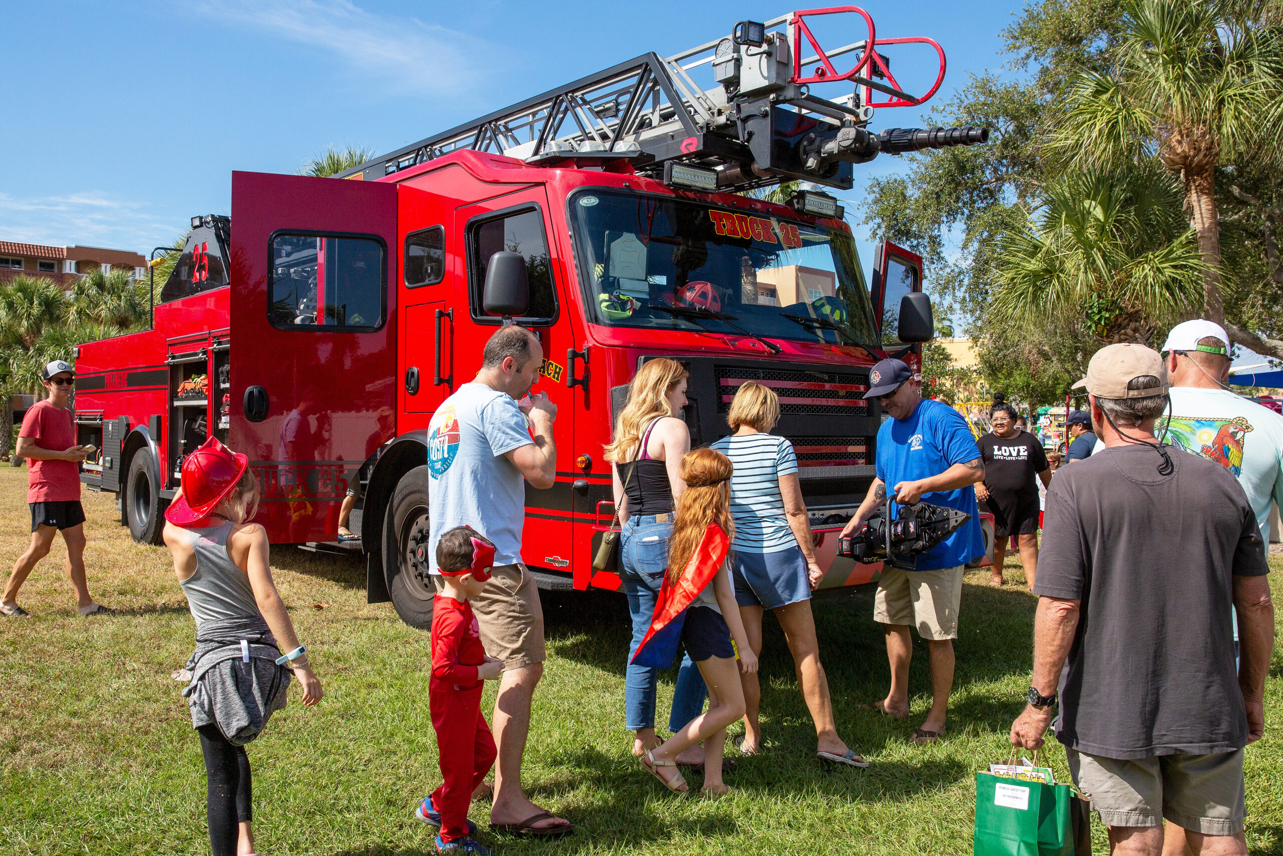 Red fire truck with extended ladder; families stand in front on a grassy lawn under a blue sky.