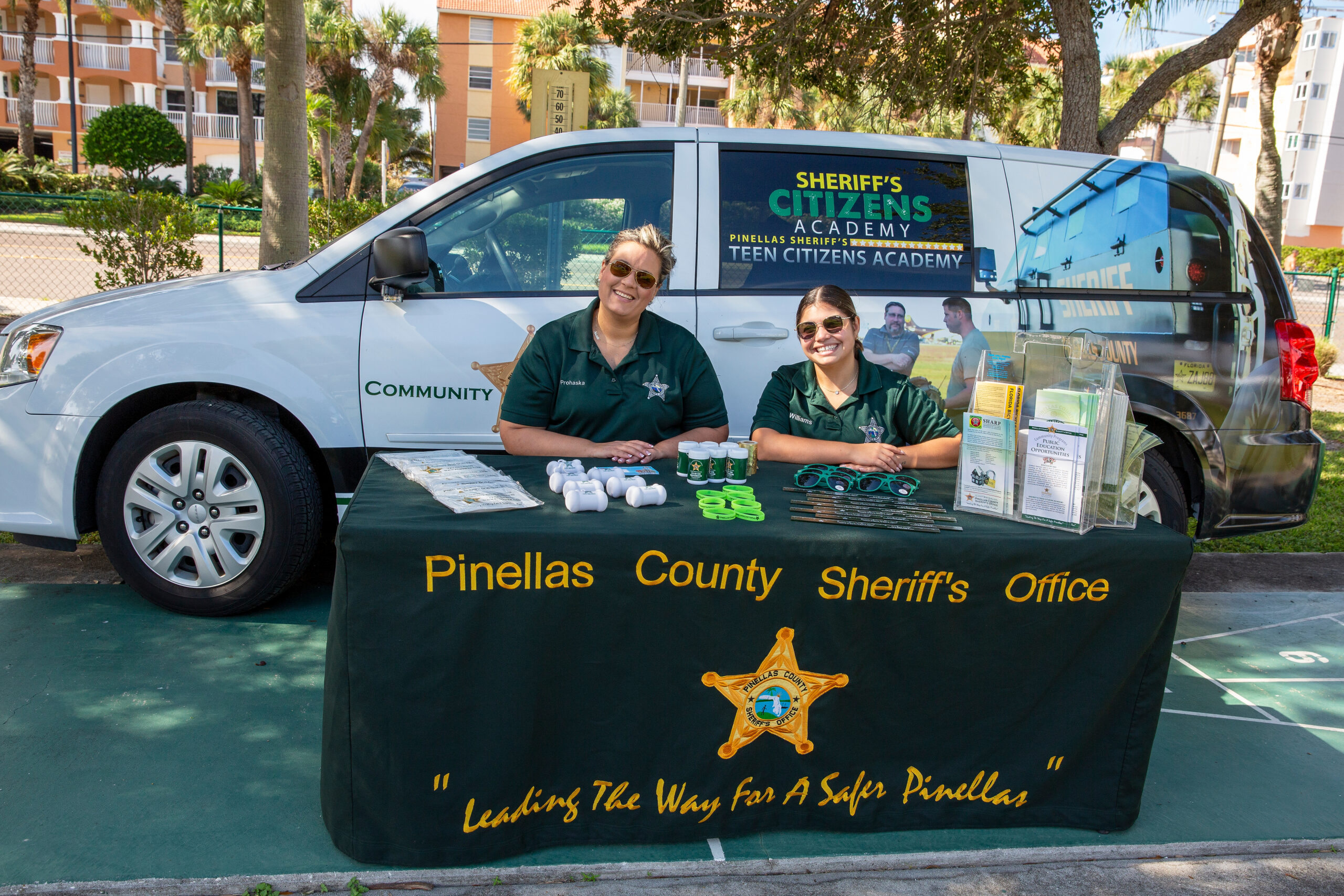Two staff members in green shirts sit at a community table in front of a Pinellas County Sheriff van labeled Citizens Academy.