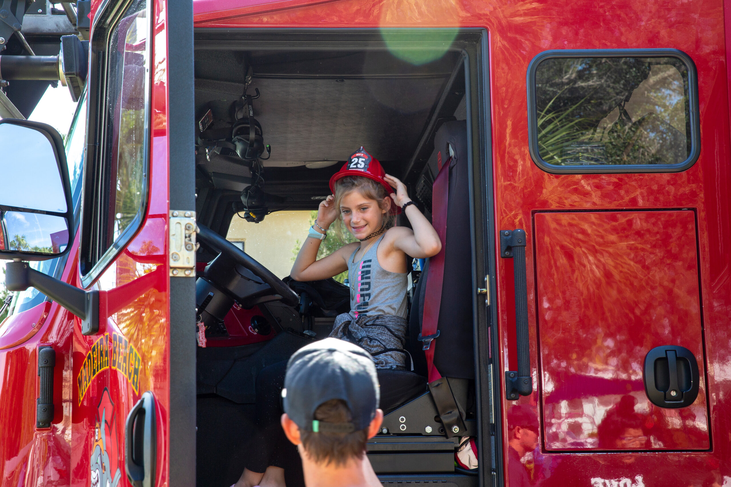 Child in a red fire helmet sits inside open fire truck doorway, smiling and holding headset/helmet.