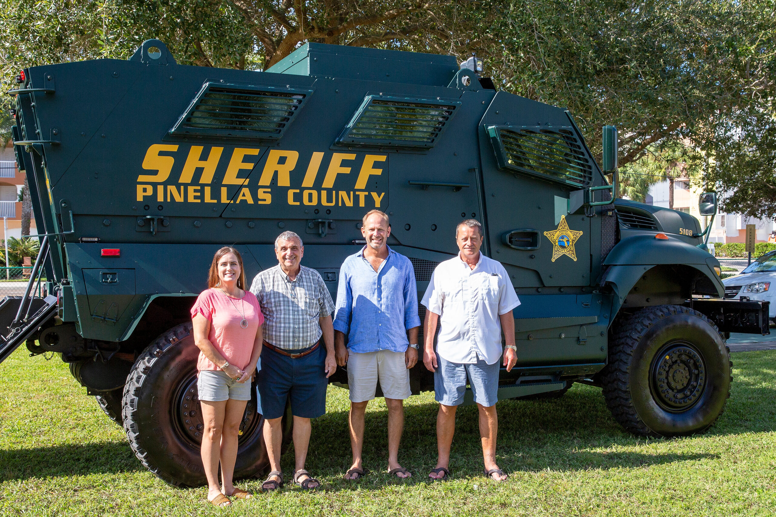 Four people stand on grass in front of a dark green vehicle with “SHERIFF PINELLAS COUNTY” lettering.