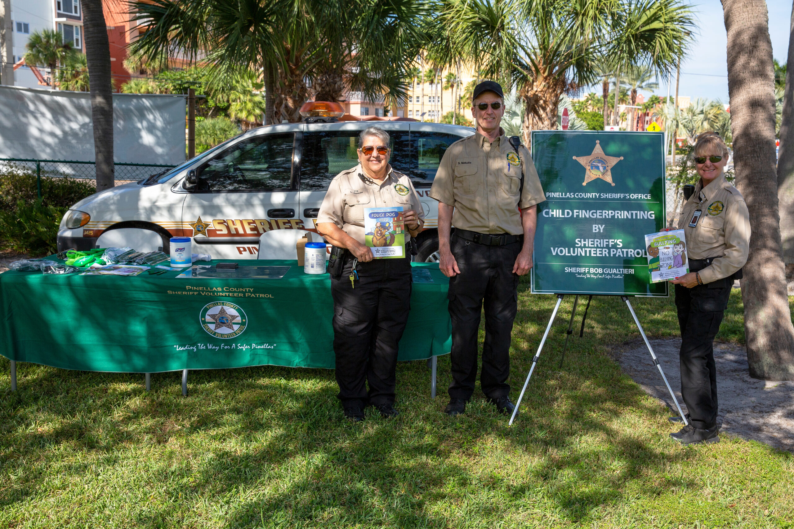 Two sheriff volunteers stand at a table with child fingerprinting sign; marked sheriff vehicle behind them; palm trees outside.