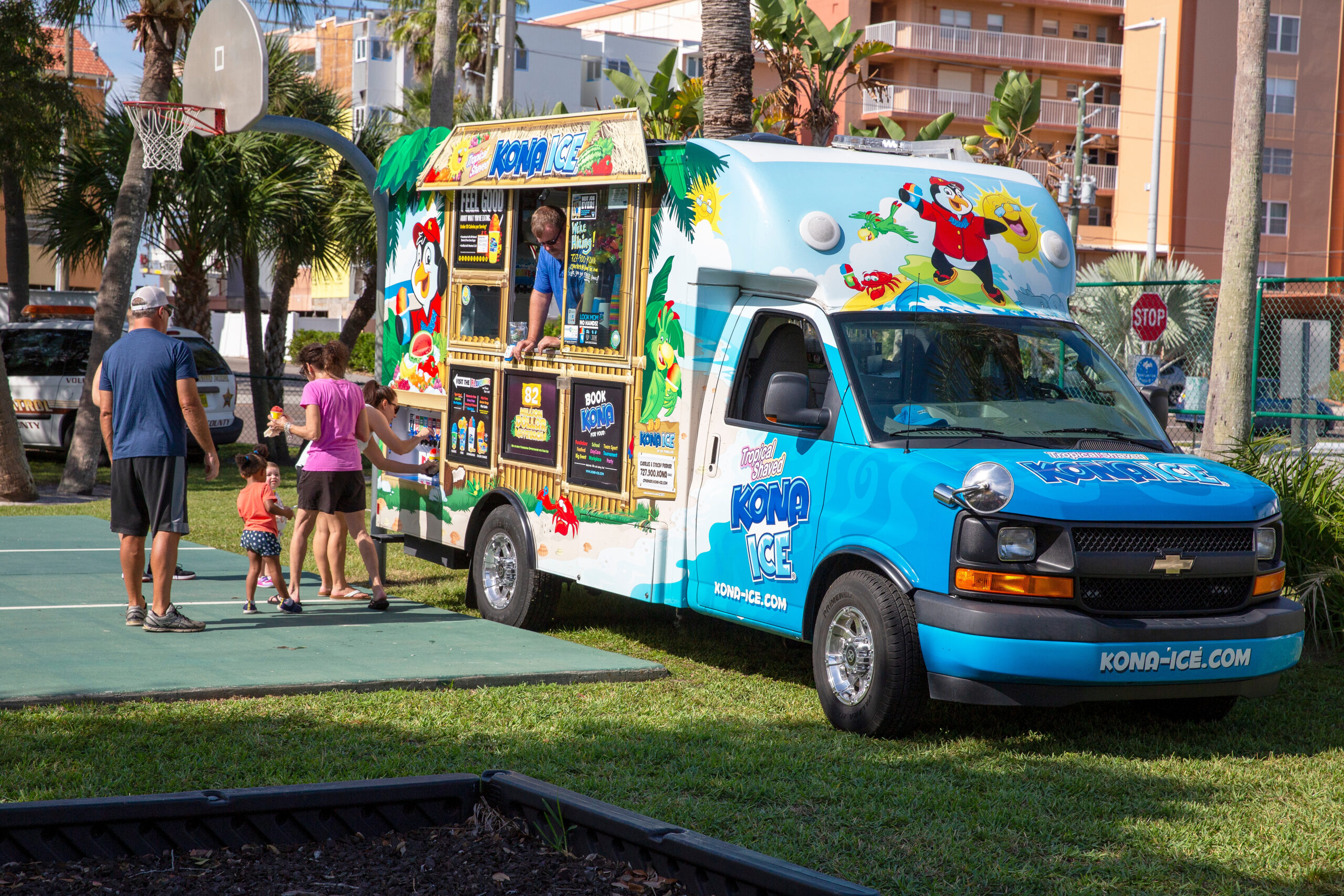 Blue Kona Ice truck with cartoon graphics parked outside; a vendor and families stand near the serving window.